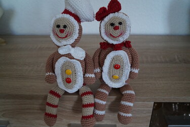 Zwei gehäkelte Lebkuchenfiguren auf einer Holzfläche, eine mit weißer Kopfbedeckung, die andere mit roter Schleife