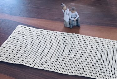 Beigefarbener gehäkelter Tischläufer mit Gitter-Filetmuster auf Holztisch, im Hintergrund zwei kleine Dekofiguren.