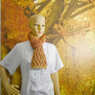Gestrickter Kurzschal mit Wellenmuster in Orange- und Beigetönen an einer Schaufensterpuppe vor herbstlichem Hintergrund