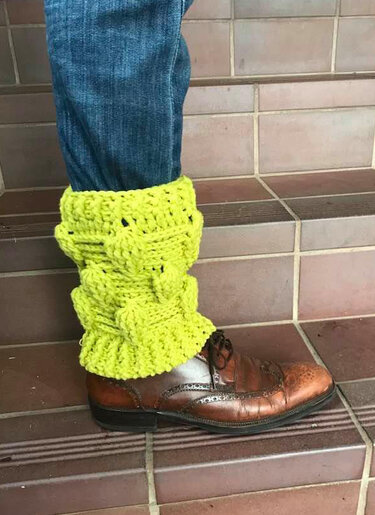 Nahaufnahme limettengrüner, gehäkelter Beinstulpen über Jeans und braunen Schuhen auf einer gefliesten Treppe.