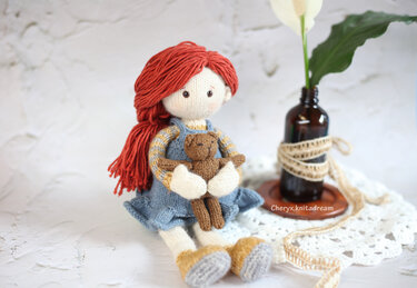 Hand-knit amigurumi girl with red yarn hair in blue dress holding small brown teddy seated on a white doily
