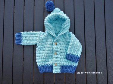 Hellblaue und dunkelblaue gehäkelte Babyjacke mit Kapuze, Bommel und drei Holzknöpfen, flach auf dunklem Holz gelegen.