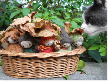 Körbchen mit fünf gehäkelten Amigurumi-Wichteln unter Blättern, rechts eine schnüffelnde Katze