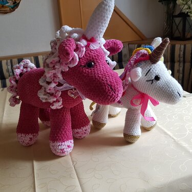 Zwei gehäkelte Amigurumi-Einhörner: ein großes pinkes mit weißem Horn und ein kleines weißes mit bunter Mähne und rosa Schleife.