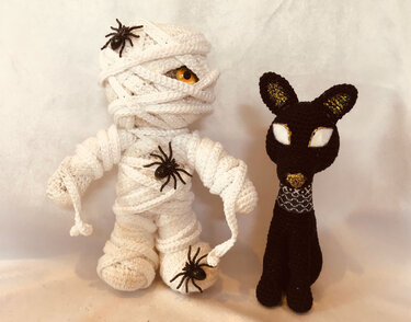 Gehäkelte Mumie mit orangefarbenen Augen und drei Spielzeugspinnen neben einer schwarzen Katze mit glitzernden Ohren