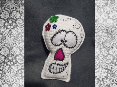 Weißer, gehäkelter Totenkopf mit schwarzen Sticknähten, rosa Filznase und bunten Blumenapplikationen vor dunklem Hintergrund.