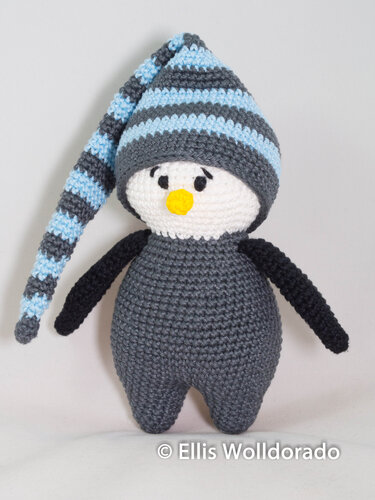 Gehäkelter Amigurumi-Pinguin, dunkelgrauer Körper, weißes Gesicht, gelber Schnabel und blau-grau gestreifte lange Mütze.