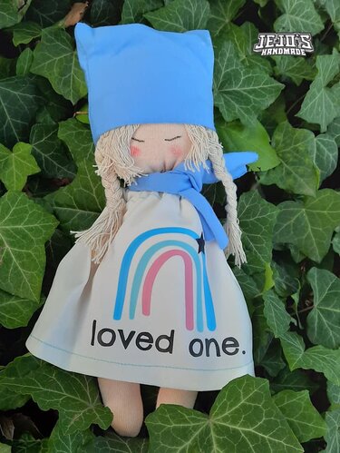 Handgefertigte Stoffpuppe mit blonden Zöpfen, blauer Mütze und Schal, weißes Kleid mit Regenbogenmotiv und Schriftzug "loved one.", liegend auf Efeu.