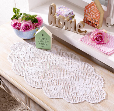 Weißer gehäkelter Tischläufer auf einer hellen Holzkommode, dekoriert mit rosa Rosen, einem Holzschriftzug und kleinen Dekoobjekten.