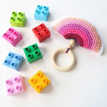 Gehäkelter Regenbogen-Greifling mit Holzring und bunten Bausteinen auf hellem Untergrund.