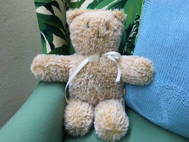 Plüschiger hellbrauner Teddy mit weißer Schleife auf grünem Sessel, blaues Strickkissen daneben