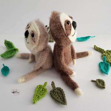 Zwei gehäkelte Amigurumi-Faultiere, hell und dunkel, sitzen Rücken an Rücken auf weißem Hintergrund mit gehäkelten grünen Blättern.