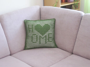 Grünes gehäkeltes Kissen mit HOME-Schriftzug und Herz auf hellem Sofa