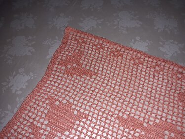Korallfarbener gehäkelter Tischläufer mit offenem Netzmuster und Bordüre auf einer hellen, floralen Tischdecke.