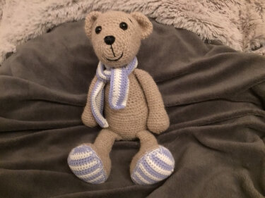 Beiger gehäkelter Amigurumi-Teddybär mit blau-weiß gestreiftem Schal sitzt auf Decke