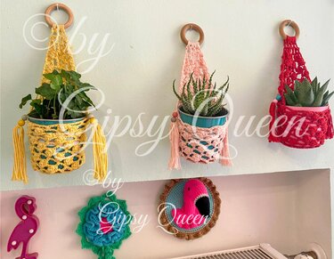 Drei gehäkelte Pflanzenampeln in Gelb, Rosa und Rot mit Holzringen und Perlen halten grüne Zimmerpflanzen an einer Wand.