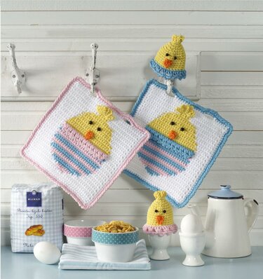 Gehäkelte Topflappen mit Kükenmotiv und passende Eierwärmer in Pastellfarben an Haken und auf einer Küchentheke.