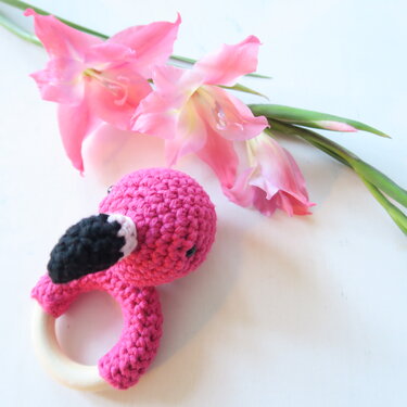 Gehäkelter pinker Flamingo-Beißring mit Holzring neben rosa Gladiolen auf weißem Hintergrund