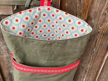 Aufhängbare olivgrüne Stoff-Wandtasche mit Außenfach, rotem Aufhängeband und innenem Sternenmuster vor einer Holzoberfläche