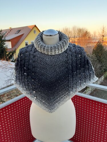 Gehäkelter Damenponcho in Grau-Ombré mit Rollkragen auf Schaufensterpuppe auf einem Balkon.