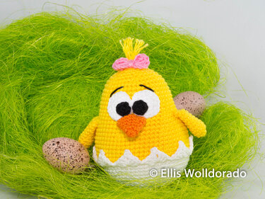 Gelbes Amigurumi-Küken mit rosa Schleife in weißer Eierschale auf grünem Dekogras, mit zwei gesprenkelten Ostereiern.