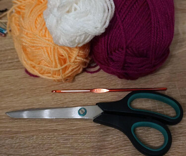 Drei Garnknäuel (weiß, apricot, magenta) mit Häkelnadel 4,0 mm und Schere auf Holzuntergrund