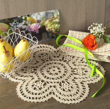 Beiges gehäkeltes Deckchen mit filigranem Blumenmuster auf Holztisch, neben Birnen und Blumen.