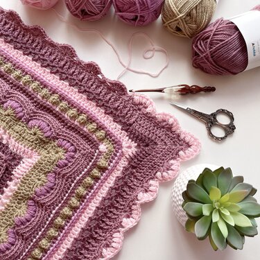 Close-up of crochet stitch textures on mauve, pink and beige baby blanket corner with yarn balls, crochet hook and decorative scissors