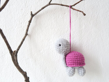 Gehäkelte Amigurumi-Schildkröte in Grau mit pinkem Panzer, an einem Ast hängend vor weißer Wand.