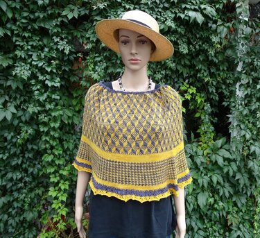 Gestrickter Damenponcho in Gelb und Anthrazit mit geometrischem Muster an Schaufensterpuppe vor Efeu