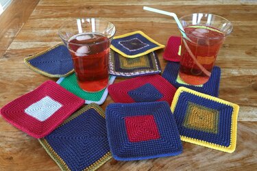 Mehrere bunte, quadratische gehäkelte Untersetzer aus Garn auf einem Holztisch, zwei Gläser mit rotem Getränk darauf