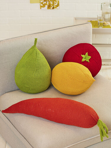 Vier gestrickte Kissen in Obst- und Gemüseform auf einem hellgrauen Sofa: grüne Birne, gelbe Zitrone, rotes Tomatenkissen und rotes Chili-Kissen