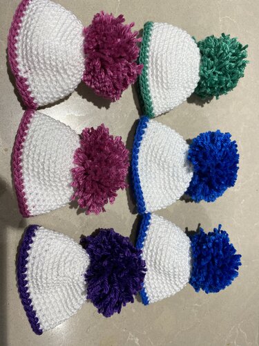 Sechs weiße gehäkelte Eierwärmer mit bunten Rändern und Pompons (Rosa, Grün, Blau, Lila) auf hellem Untergrund.