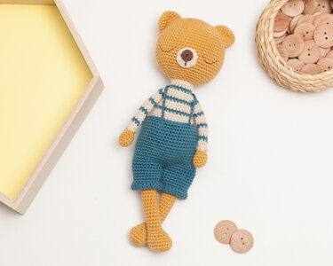 Mustard-colored crocheted amigurumi teddy wearing cream-and-teal striped sweater and teal overalls, flat on white background with wooden buttons