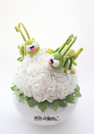 Zwei grüne gehäkelte Amigurumi-Insekten mit großen Augen auf einem Bouquet weißer Rosen in einem weißen Topf, umgeben von perlenähnlicher Dekoration.