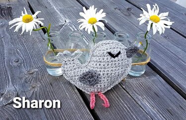 Gehäkeltes graues Amigurumi-Vögelchen mit rosa Füßen auf Holz, neben Glasflaschen mit Gänseblümchen.