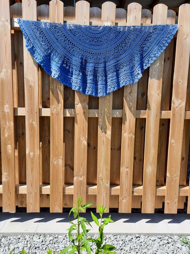 Blue crocheted crescent shawl with lace stitches draped on a wooden fence.