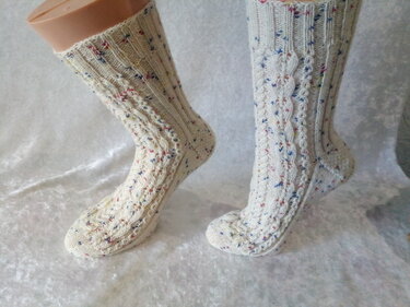Gestrickte cremefarbene Socken mit Zopfmuster und bunten Sprenkeln auf hellem Hintergrund.