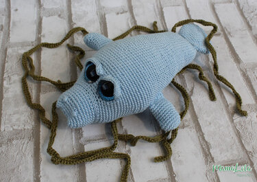 Hellblaues gehäkeltes Seekuh-Amigurumi mit großen glänzenden Augen und dunkelgrünen Häkelranken auf hellem Ziegelmuster.