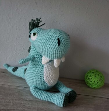 Gehäkelter mintgrüner Amigurumi-Dino mit weißem Bauch und dunkelgrünen Punkten, sitzend auf Holzfläche