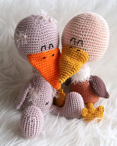 Zwei gehäkelte Amigurumi-Vögel in Pastellfarben mit berührenden Schnäbeln auf weißem, flauschigem Untergrund.
