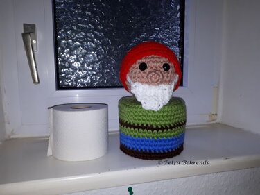 Gehäkelter Wichtel mit roter Mütze und weißem Bart neben einer Toilettenpapierrolle auf einer Fensterbank.