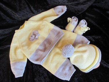 Gestricktes Baby-Set: Jacke, Hose, Bommelmütze und Babyschuhe in Gelb und Grau auf dunklem Hintergrund.