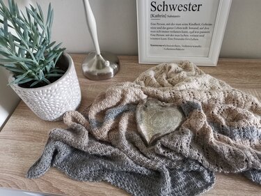 Gestricktes Dreieckstuch in Beige- und Grautönen mit feinem Lochmuster, dekorativ auf einem Holztisch drapiert neben einer Zimmerpflanze und einem Herzornament.