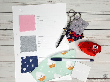 Nahaufnahme eines Stoffpasses mit rosa-, grau- und blaugemusterten Stoffmustern, umgeben von einer Schere, einem Kleberoller, einem Stift und weiteren Stoffresten auf einem Holztisch.