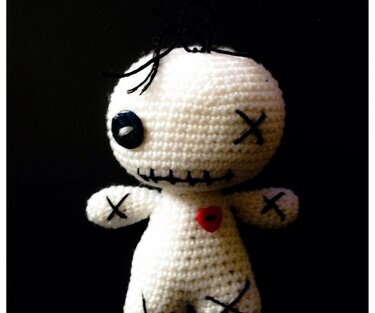 Weiße gehäkelte Amigurumi-Puppe mit schwarzem Knopfauge, einem gestickten X als weiteres Auge, gesticktem Mund, schwarzem Haar und rotem Herzknopf vor schwarzem Hintergrund.
