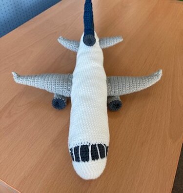 Gehäkeltes Amigurumi‑Flugzeug in Weiß und Grau auf Holztisch, Draufsicht