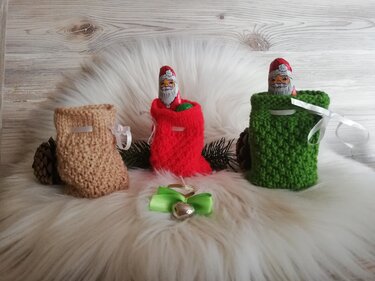 Drei gestrickte Weihnachtssäckchen (Beige, Rot, Grün) mit Schoko-Nikoläusen auf weißem Fell vor Holzpaneel