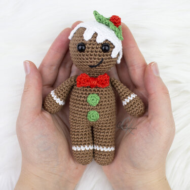 Gehäkeltes Amigurumi-Lebkuchenmännchen mit weißen Zuckerguss-Details, roter Fliege und grünen Knöpfen, in zwei Händen gehalten
