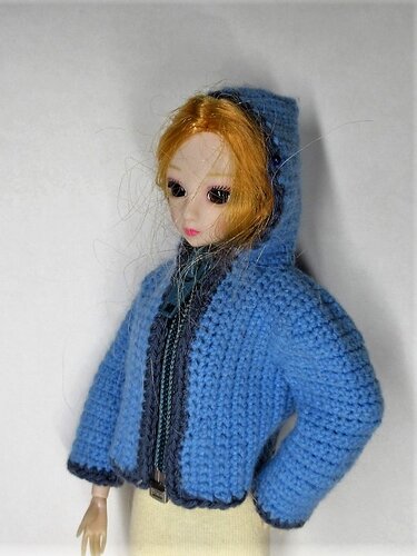 Puppe trägt hellblaue gehäkelte Kapuzenjacke mit dunklem Rand, halbe Frontansicht